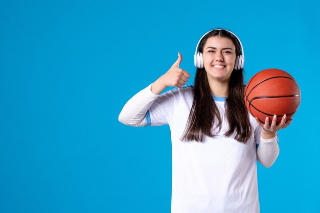 Mulher jovem com fones de ouvido segurando uma bola de basquete na parede azul de frente