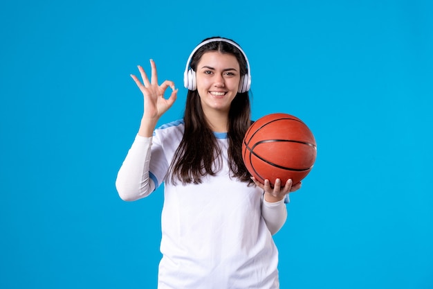 Mulher jovem com fones de ouvido segurando uma bola de basquete na parede azul de frente