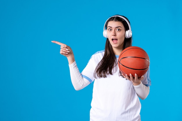 Mulher jovem com fones de ouvido segurando uma bola de basquete na parede azul de frente