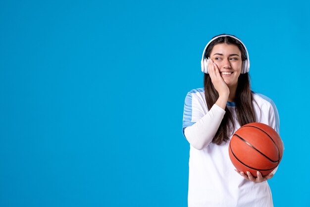 Mulher jovem com fones de ouvido segurando uma bola de basquete na parede azul de frente