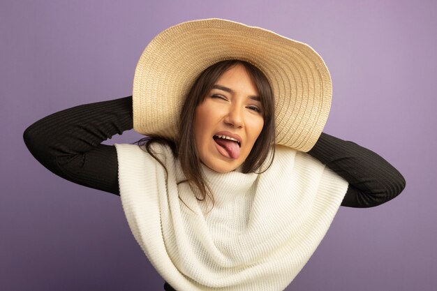 Mulher jovem com lenço branco e chapéu de verão olhando para a frente feliz e alegre, mostrando a língua em pé sobre a parede roxa