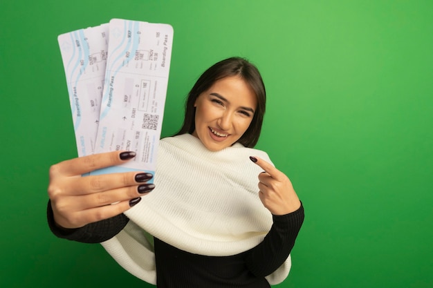 Mulher jovem com lenço branco segurando passagens aéreas e apontando com figner para eles sorrindo alegremente