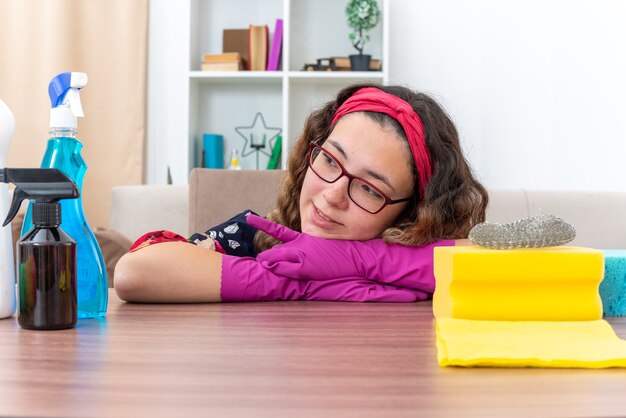 Mulher jovem com luvas de borracha, olhando para o lado, sorrindo feliz e positiva, sentada à mesa com material de limpeza e ferramentas na luz da sala de estar