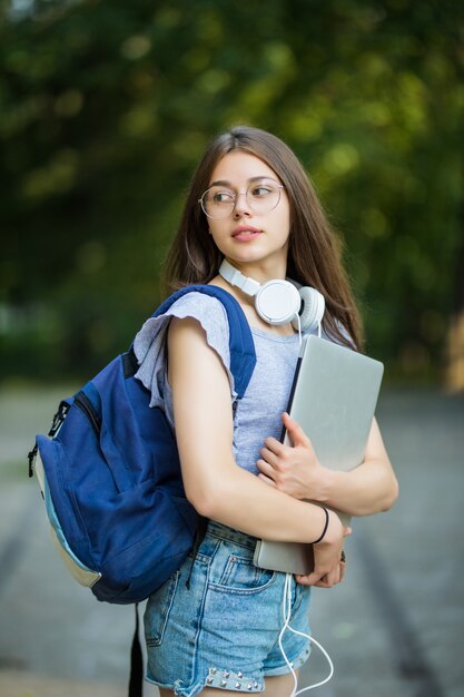 Mulher jovem com mochila caminhando pelo parque verde com laptop prateado nas mãos