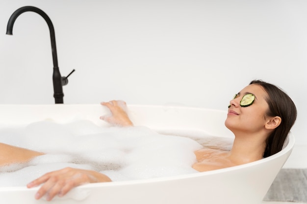 Mulher jovem com pepino nos olhos relaxando na banheira cheia de água e espuma