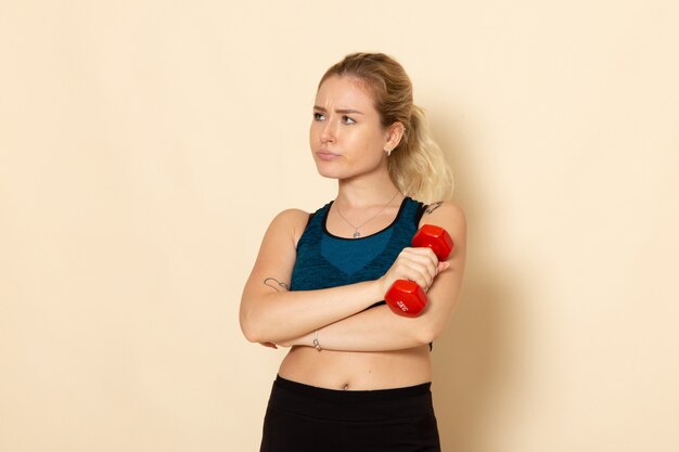 Mulher jovem com roupa esportiva, vista frontal, segurando halteres vermelhos na parede branca, saúde, esporte, corpo, beleza, treino