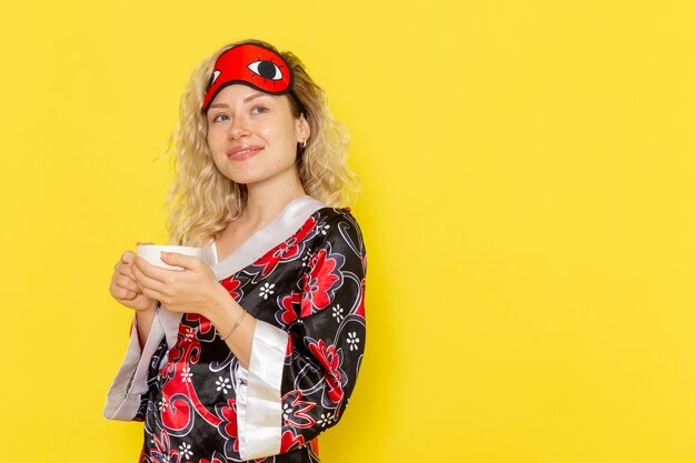 Mulher jovem com roupão de noite e máscara de olho se preparando para dormir, bebendo café na cama de noite modelo feminino de sono de parede amarela