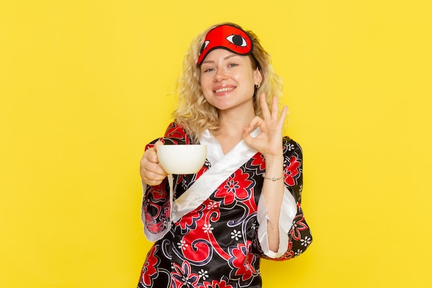 Mulher jovem com roupão de noite e máscara de olho se preparando para dormir, segurando uma xícara de café, sorrindo na parede amarela.