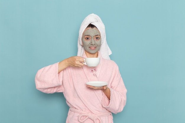 Mulher jovem com roupão rosa após o banho, bebendo café com um sorriso na parede azul, beleza limpeza água limpa creme para cuidar