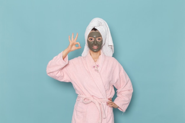 Mulher jovem com roupão rosa após o banho com máscara na parede azul beleza água creme autocuidado chuveiro banheiro