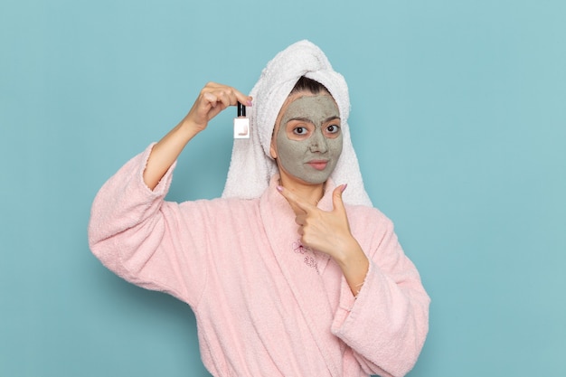 Mulher jovem com roupão rosa após o banho segurando esmalte de unha na parede azul beleza limpeza água limpa creme autocuidado chuveiro