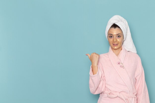 Mulher jovem com roupão rosa após o banho sorrindo na parede azul beleza água creme autocuidado chuveiro banheiro