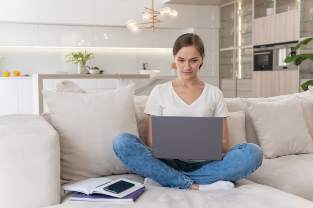 Mulher jovem com roupas casuais trabalhando em um laptop enquanto está sentada no sofá em casa