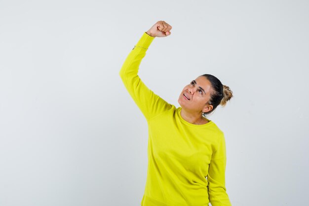 Mulher jovem com suéter amarelo e calça preta cerrando os punhos e parecendo feliz