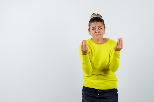 Mulher jovem com suéter amarelo e calça preta, mostrando um gesto italiano e parecendo impressionada