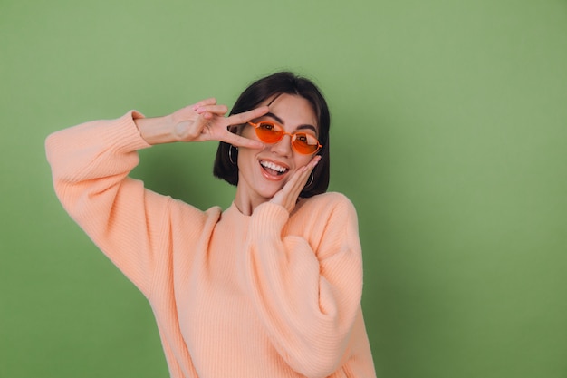 Mulher jovem com suéter casual cor de pêssego e óculos laranja isolados na parede verde-oliva engraçada fazendo o gesto de vitória copiar o espaço