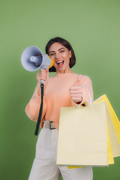 Mulher jovem com suéter casual cor de pêssego isolado na parede verde oliva grito no megafone segurando sacolas de compras e anuncia promoção de venda de descontos