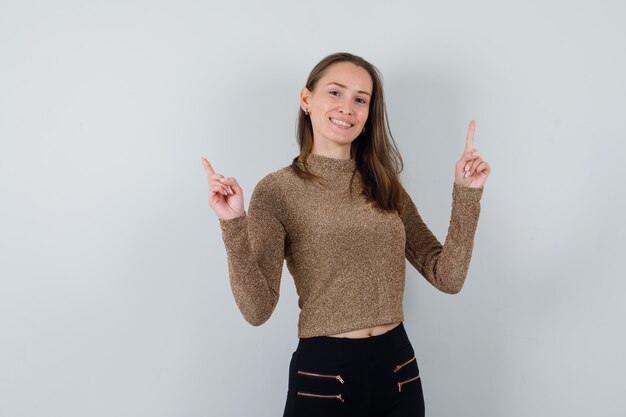 Mulher jovem com suéter dourado e calça preta apontando para cima com os dois dedos indicadores e parecendo feliz