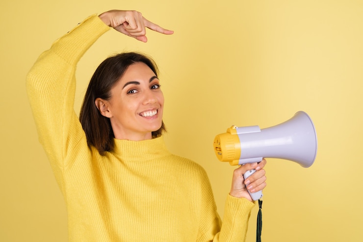 Foto grátis mulher jovem com suéter quente amarelo com alto-falante de megafone e dedo apontando para a direita