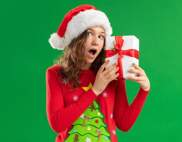 Mulher jovem com suéter vermelho de Natal e chapéu de Papai Noel segurando um presente e fica surpresa em pé sobre um fundo verde