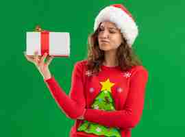 Foto grátis mulher jovem com suéter vermelho de natal e chapéu de papai noel segurando um presente olhando para ele com expressão cética em pé sobre fundo verde