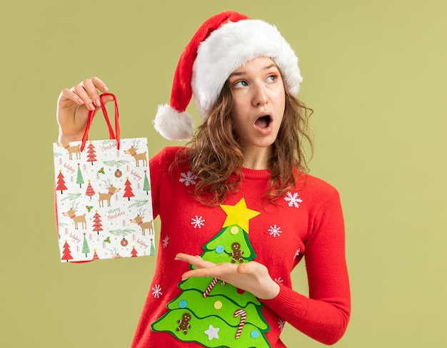 Foto grátis mulher jovem com suéter vermelho de natal e chapéu de papai noel segurando um saco de papel com presentes de natal e apresentando um braço parecendo surpresa em pé sobre a parede verde