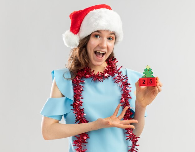 Mulher jovem com top azul e chapéu de Papai Noel com enfeites em volta do pescoço segurando cubos de brinquedo com data de Natal se apresentando com o braço da mão feliz e animada