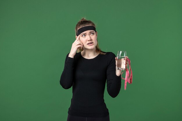 Mulher jovem com um copo de água e medida de cintura em fundo verde saúde esporte corpo preto malhação atleta de frente