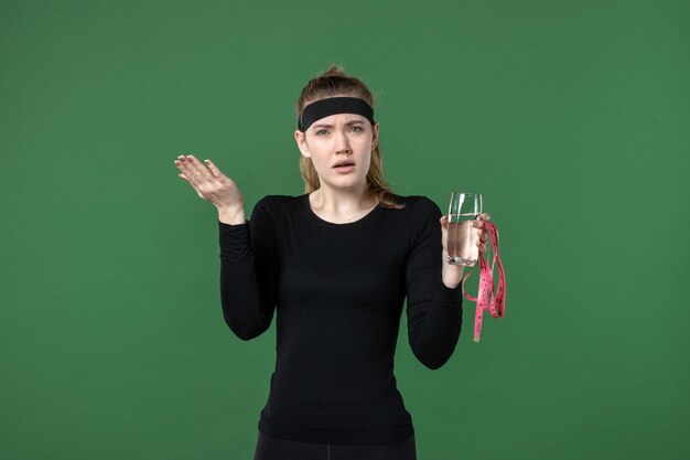 Mulher jovem com um copo de água e medida de cintura em fundo verde saúde esporte corpo preto treino mulher atleta de frente