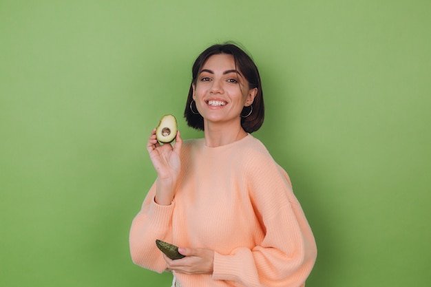 Mulher jovem com um suéter casual cor de pêssego, isolado em uma parede verde oliva, segurando um abacate, um conceito de saúde e cuidados com a pele, copie o espaço