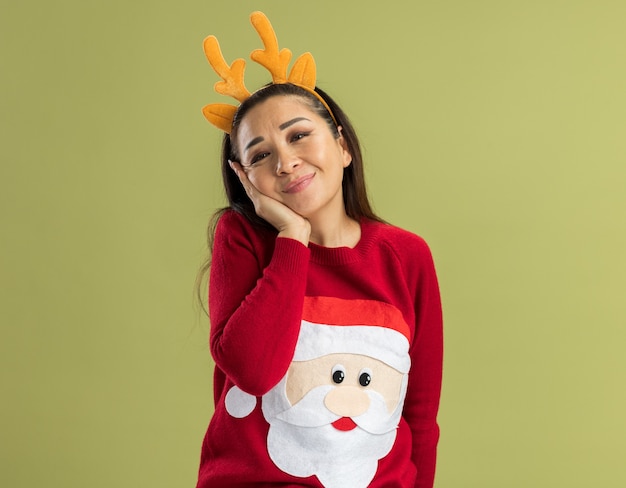 Mulher jovem com um suéter vermelho de natal, vestindo uma borda engraçada com chifres de veado e um sorriso feliz e positivo