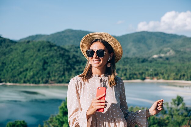 Mulher jovem com um vestido fofo de verão, chapéu de palha e óculos escuros dançando com o smartphone disponível