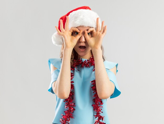 Mulher jovem com uma blusa azul e um chapéu de Papai Noel com enfeites em volta do pescoço olhando por entre os dedos fazendo um gesto binocular sendo surpreendida