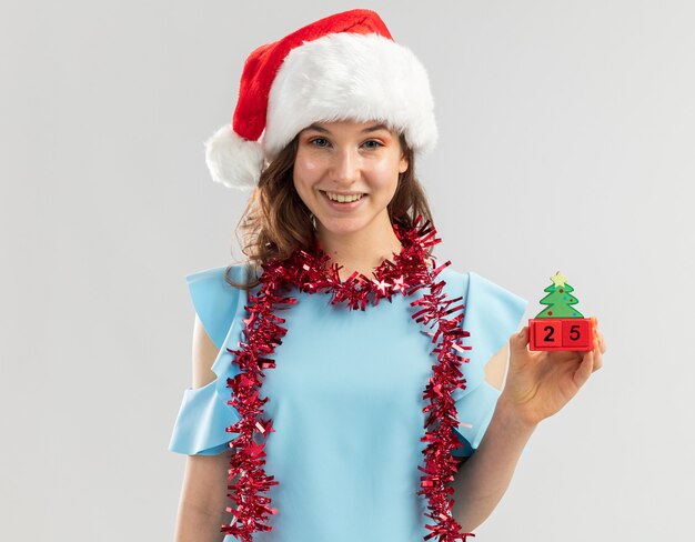 Mulher jovem com uma blusa azul e um chapéu de Papai Noel com enfeites em volta do pescoço segurando cubos de brinquedo com data de Natal e sorrindo alegremente