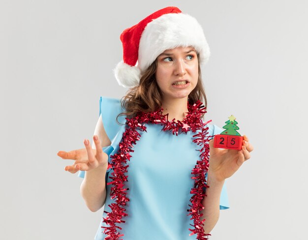 Mulher jovem com uma blusa azul e um chapéu de Papai Noel com enfeites no pescoço segurando cubos de brinquedo com a data de Natal, parecendo confusa e descontente com o braço esticado