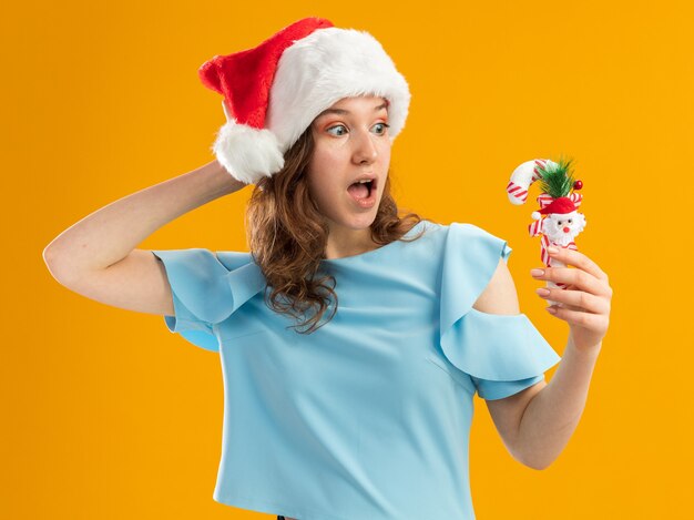 Mulher jovem com uma blusa azul e um chapéu de Papai Noel segurando uma bengala de doces de natal, olhando para ela com a mão na cabeça