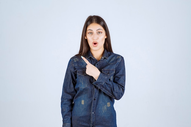 Mulher jovem com uma camisa jeans apontando para o lado esquerdo com emoções no rosto