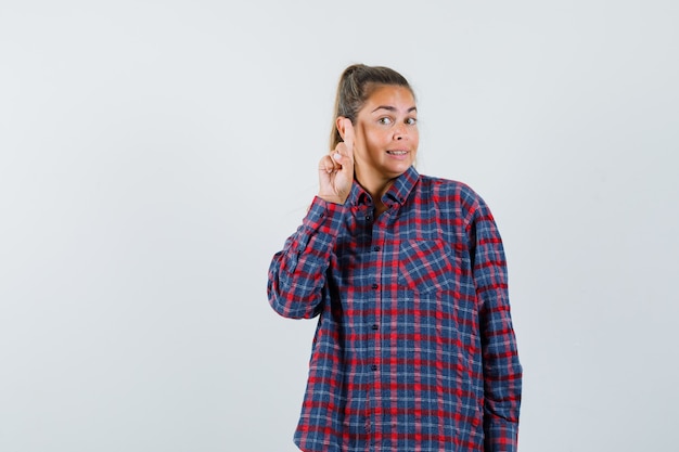 Mulher jovem com uma camisa xadrez levantando o dedo indicador num gesto de eureka e parecendo pensativa