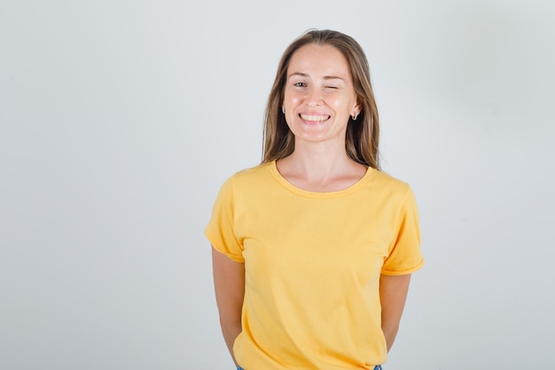 Mulher jovem com uma camiseta amarela piscando e segurando as mãos atrás dela e parecendo feliz