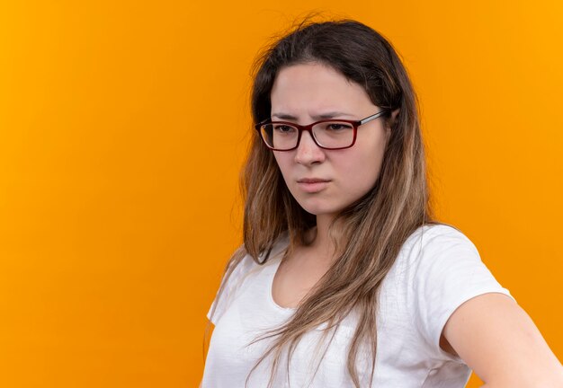 Mulher jovem com uma camiseta branca olhando para o lado com uma expressão pensativa no rosto pensando e descontente em pé na parede laranja