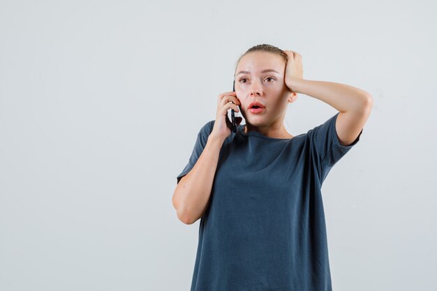 Mulher jovem com uma camiseta cinza falando no celular e parecendo chocada