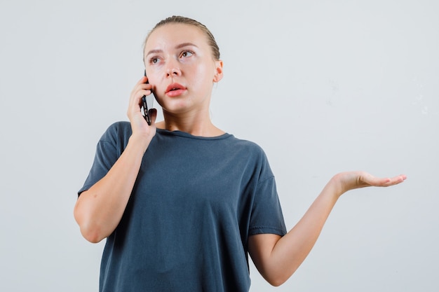 Mulher jovem com uma camiseta cinza falando no celular e parecendo confusa