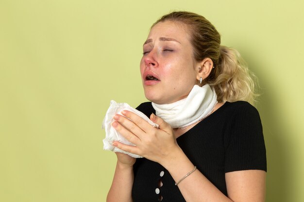 Mulher jovem com uma toalha branca em volta da garganta, de frente, sentindo-se muito enjoada e snezzing na parede verde-clara.