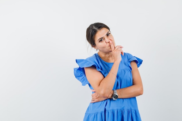 Mulher jovem com vestido azul mostrando gesto de silêncio e olhando com cuidado