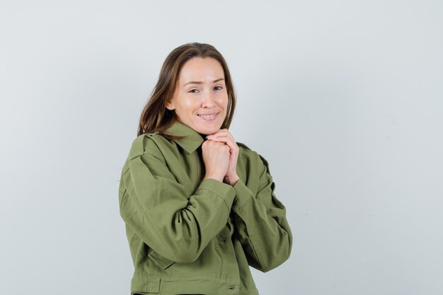 Mulher jovem combinando as mãos no peito em uma jaqueta verde e parecendo satisfeita. vista frontal.
