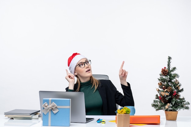 Mulher jovem confiante com chapéu de Papai Noel sentada a uma mesa com uma árvore de Natal e um presente e apontando para cima no lado esquerdo em fundo branco