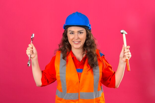 Mulher jovem Construtor em uniforme de construção e capacete de segurança em pé com chave e martelo nas mãos levantadas, sorrindo amigável sobre parede rosa isolada