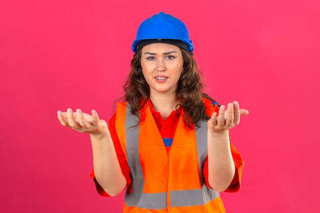 Mulher jovem construtor em uniforme de construção e capacete de segurança em pé com expressão confusa com braços e mãos levantadas sobre parede rosa isolada