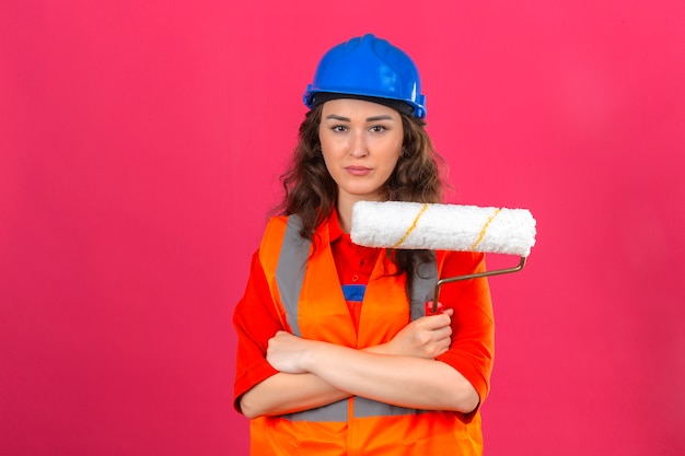 Mulher jovem Construtor em uniforme de construção e capacete de segurança em pé com os braços cruzados, segurando o rolo de pintura com cara séria sobre parede rosa isolada