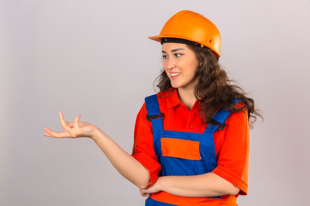 Foto grátis mulher jovem construtor em uniforme de construção e capacete de segurança, fazendo perguntas com a mão levantada em pé sobre a parede branca isolada